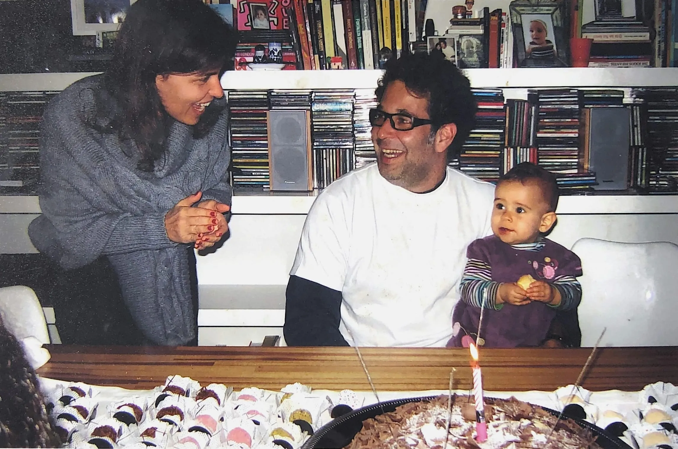 Familienfoto am Geburtstagstisch: Eine Frau und ein Mann lachen sich an, daneben sitzt ein Baby, vorne steht ein Kuchen mit Kerze.